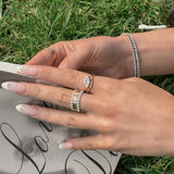 A close-up of a person's hands resting on a book outdoors on green grass, wearing a silver tennis bracelet, a silver ring with a clear center stone, and a gold ring with a stone-set band.