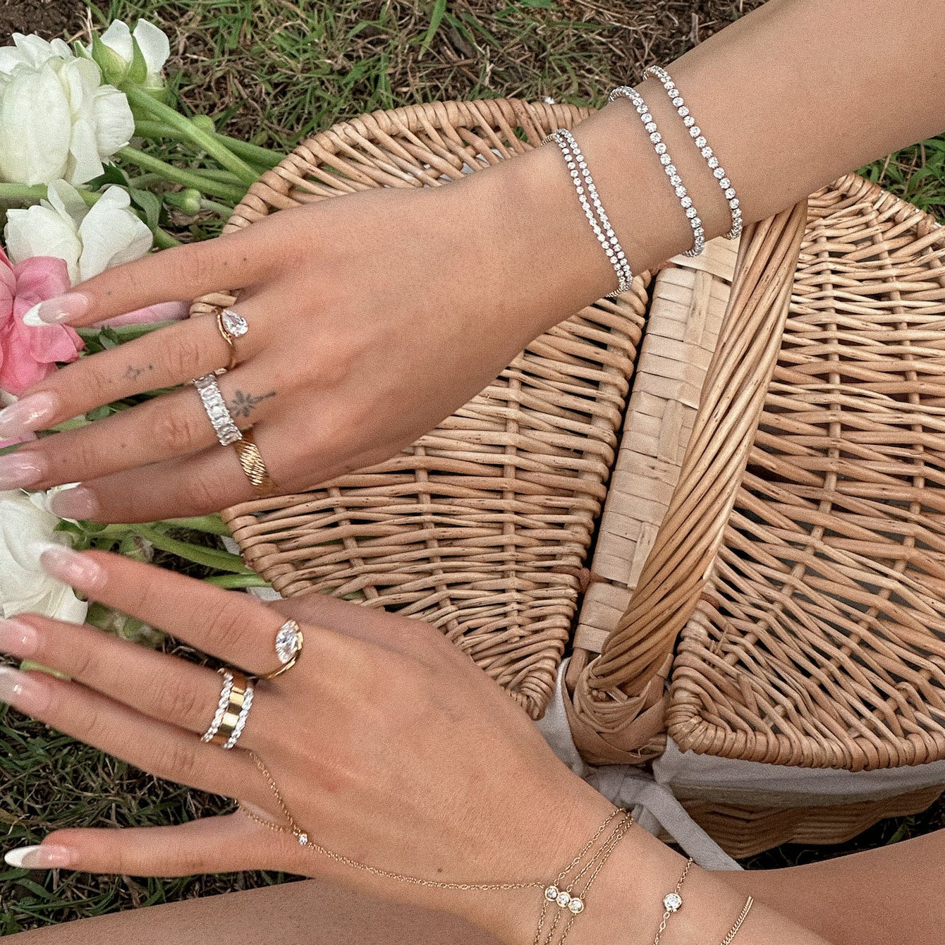 A close-up lifestyle shot of a person's hands and arms adorned with delicate silver jewelry, including rings, bracelets, and a chain necklace, resting near a wicker picnic basket on a grassy lawn with white and pink flowers.