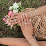 A close-up lifestyle shot of a person's hands and arms adorned with multiple delicate gold and silver rings, bracelets, and necklaces, resting on a wicker basket with pink and white flowers on a grassy lawn.