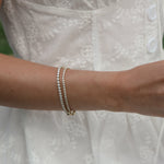 A close-up of a person in a white embroidered dress holding an open book outdoors, wearing multiple delicate rings and bracelets on both hands.