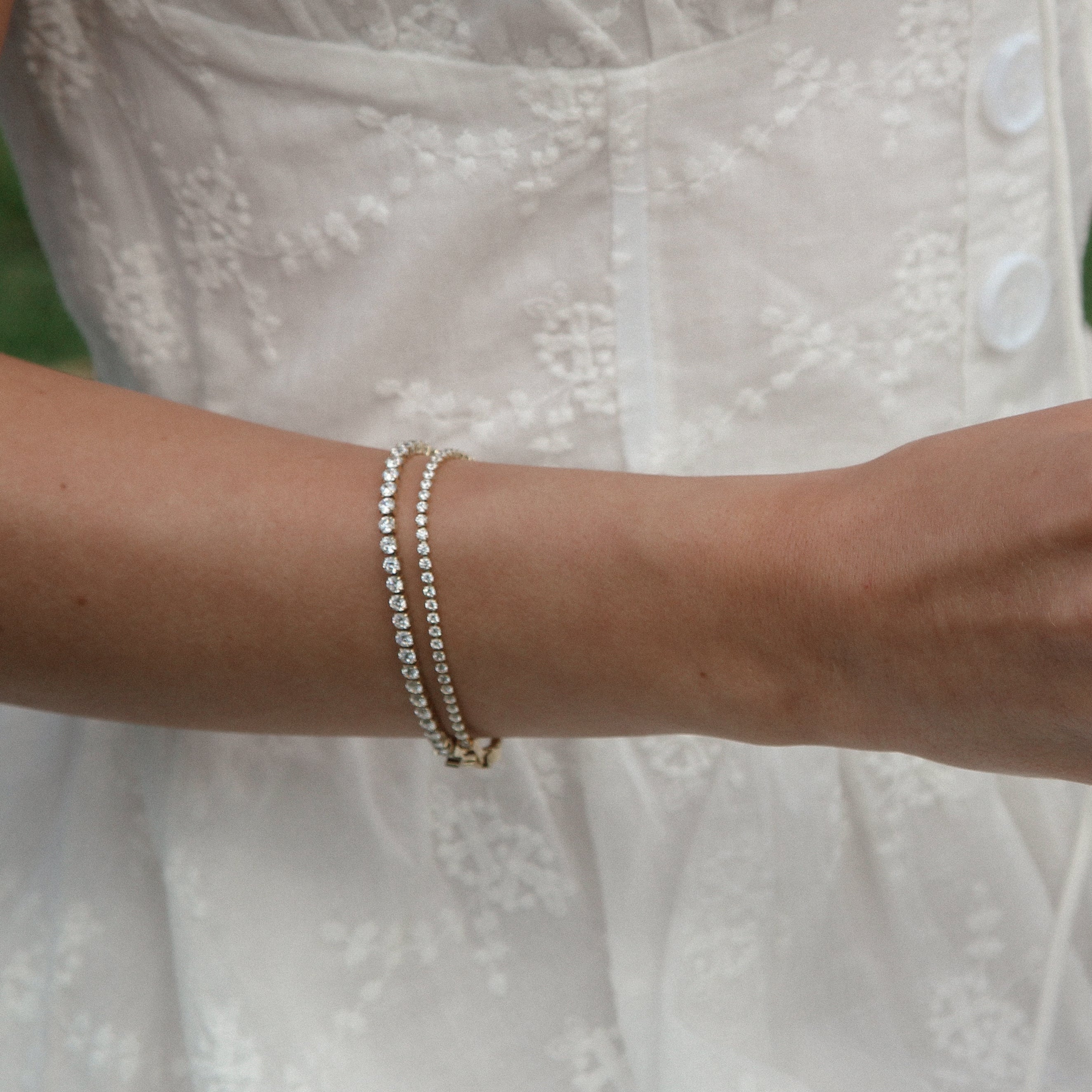 A close-up of a person in a white embroidered dress holding an open book outdoors, wearing multiple delicate rings and bracelets on both hands.