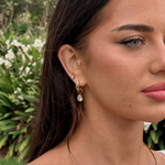 A close-up of a woman's ear wearing multiple gold hoop and stud earrings, including a dangling gold hoop with a clear teardrop gemstone, against a blurred green garden background.