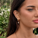 A close-up of a woman's ear wearing multiple gold hoop and stud earrings, including a dangling gold hoop with a clear teardrop gemstone, against a blurred green garden background.